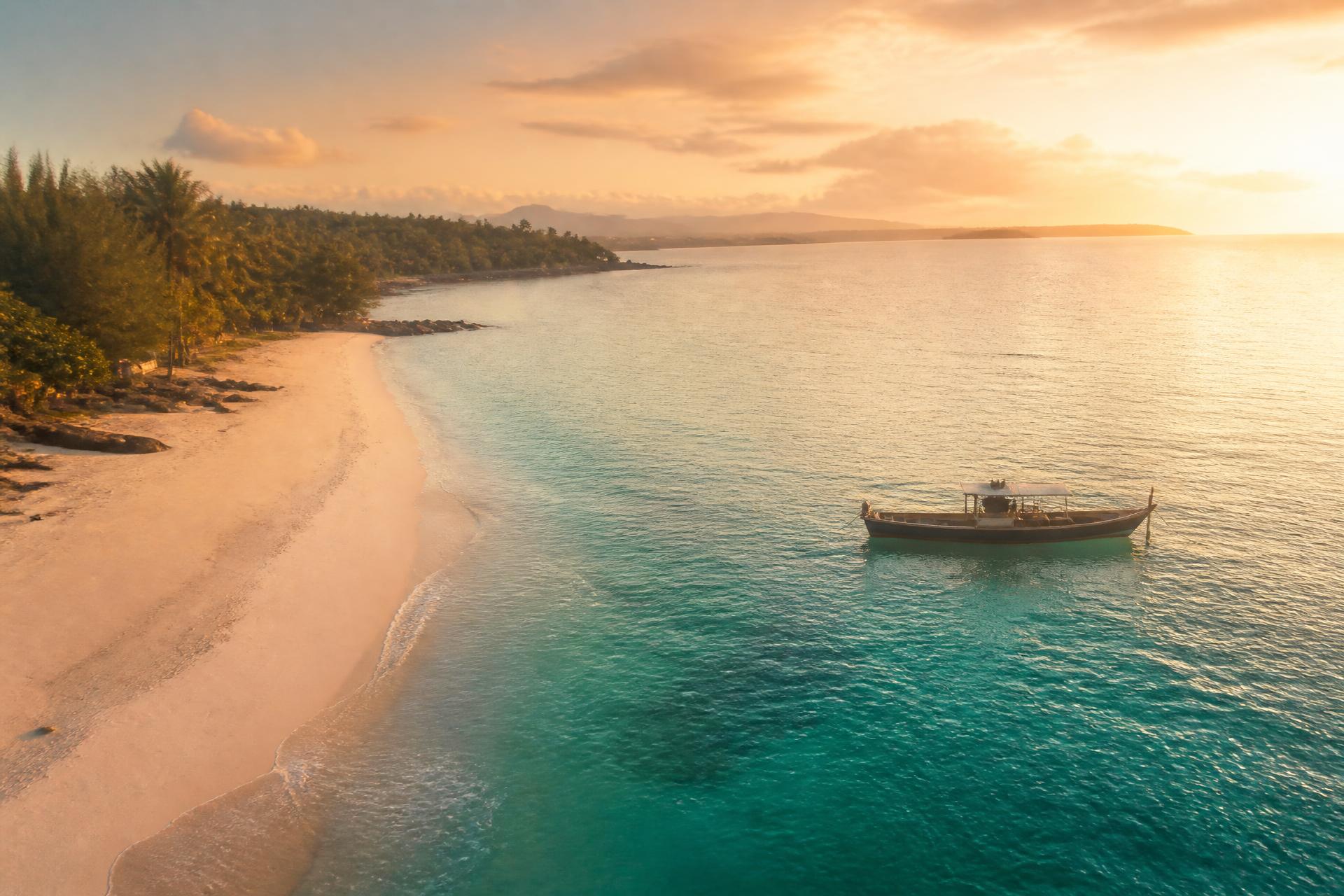 Turquoise coastline at sunrise with a wooden boat moored off a pale sand beach